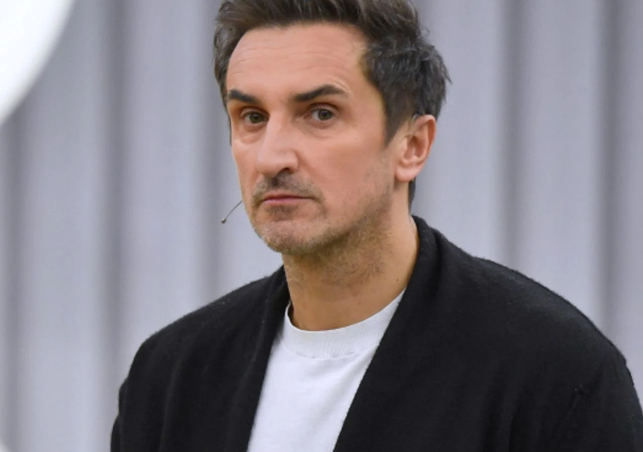 Sebastian Karpiel-Bułecka Mężczyzna o ciemnych włosach, ubrany w białą koszulkę i czarne okrycie, z mikrofonem przy twarzy, na tle jasnej, rozmytej ściany.