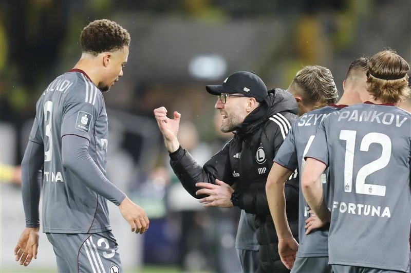 Marek Papszun i piłkarze Legii Warszawa Trener udziela wskazówek zawodnikom drużyny piłkarskiej ubranemu w szare stroje z nazwiskami na plecach, wszyscy skupieni na rozmowie podczas przerwy w meczu.