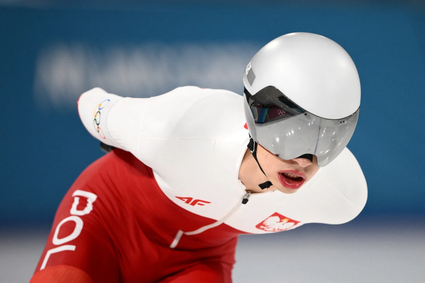 Łyżwiarz szybki w biało-czerwonym stroju z napisem POL i orłem na piersi, w aerodynamicznej pozycji podczas wyścigu, w kasku i okularach ochronnych.