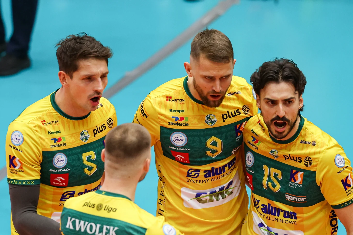 Trzech siatkarzy w żółto-zielonych koszulkach z wyraźnymi numerami i nazwiskami drużyny Aluron CMC Warta Zawiercie okazuje emocje podczas rozgrywki na hali sportowej.