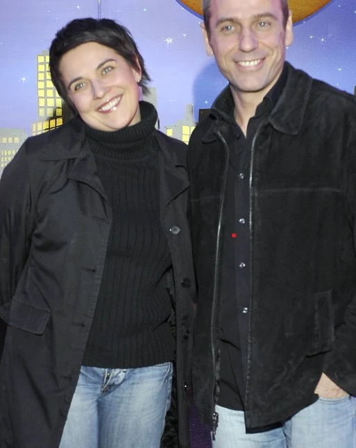 Dwoje dorosłych ubranych w ciemne ubrania stoi razem na tle bajkowej scenografii z widocznymi wysokimi budynkami i dekoracjami gwiazd nocą, uśmiechając się do aparatu.