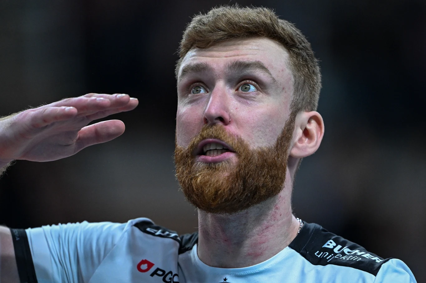 Mężczyzna z rudą brodą wykonuje gest salutowania, ubrany w sportową koszulkę, patrzy w górę w skupieniu. Tło rozmyte, uwaga skupiona na jego twarzy i wyrazie emocji.