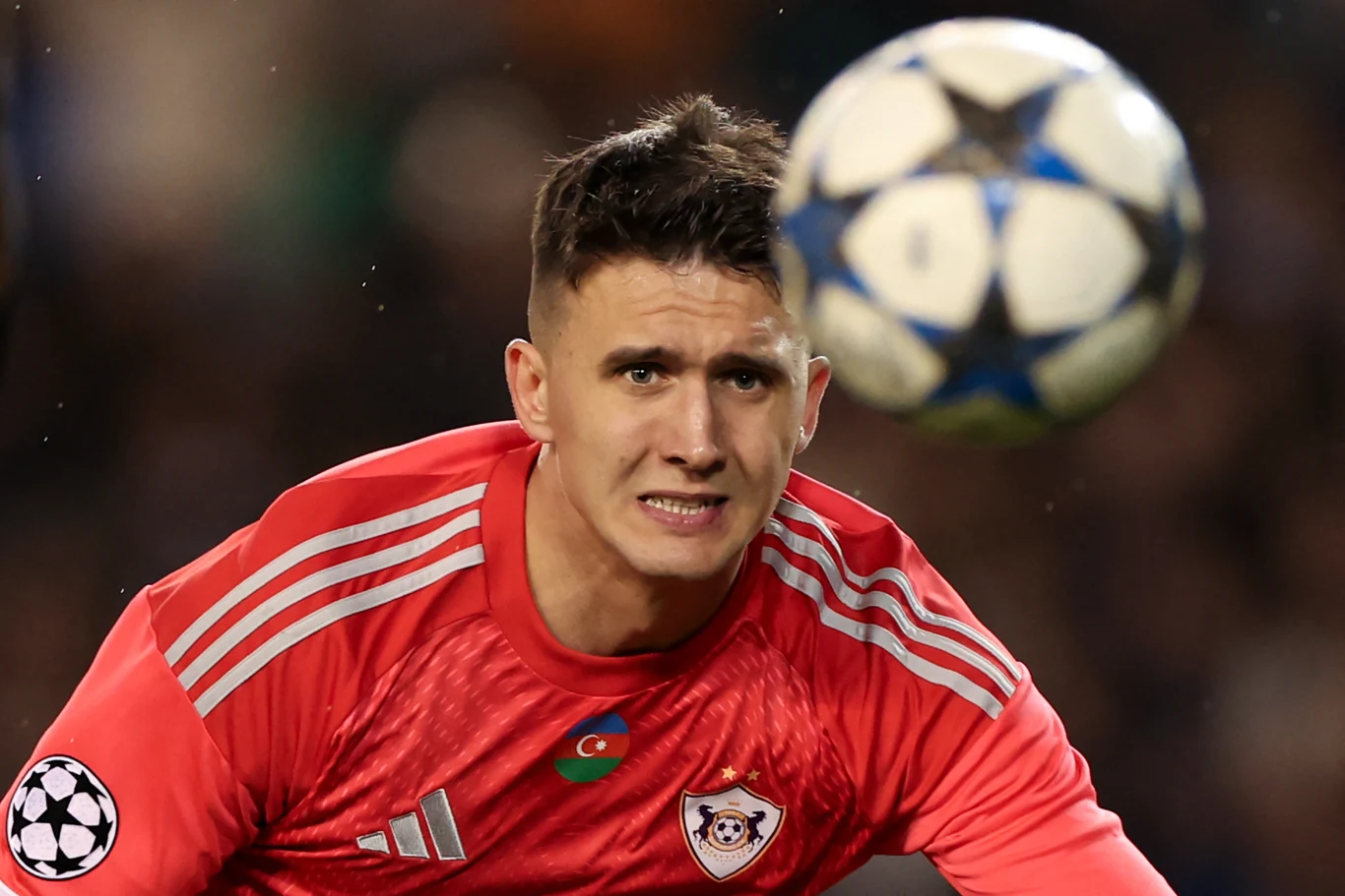 Mateusz Kochalski w barwach Karabachu Agdam Bramkarz w czerwonym stroju koncentruje się na lecącej prosto w niego piłce podczas meczu piłki nożnej, w tle rozmyte trybuny stadionu.