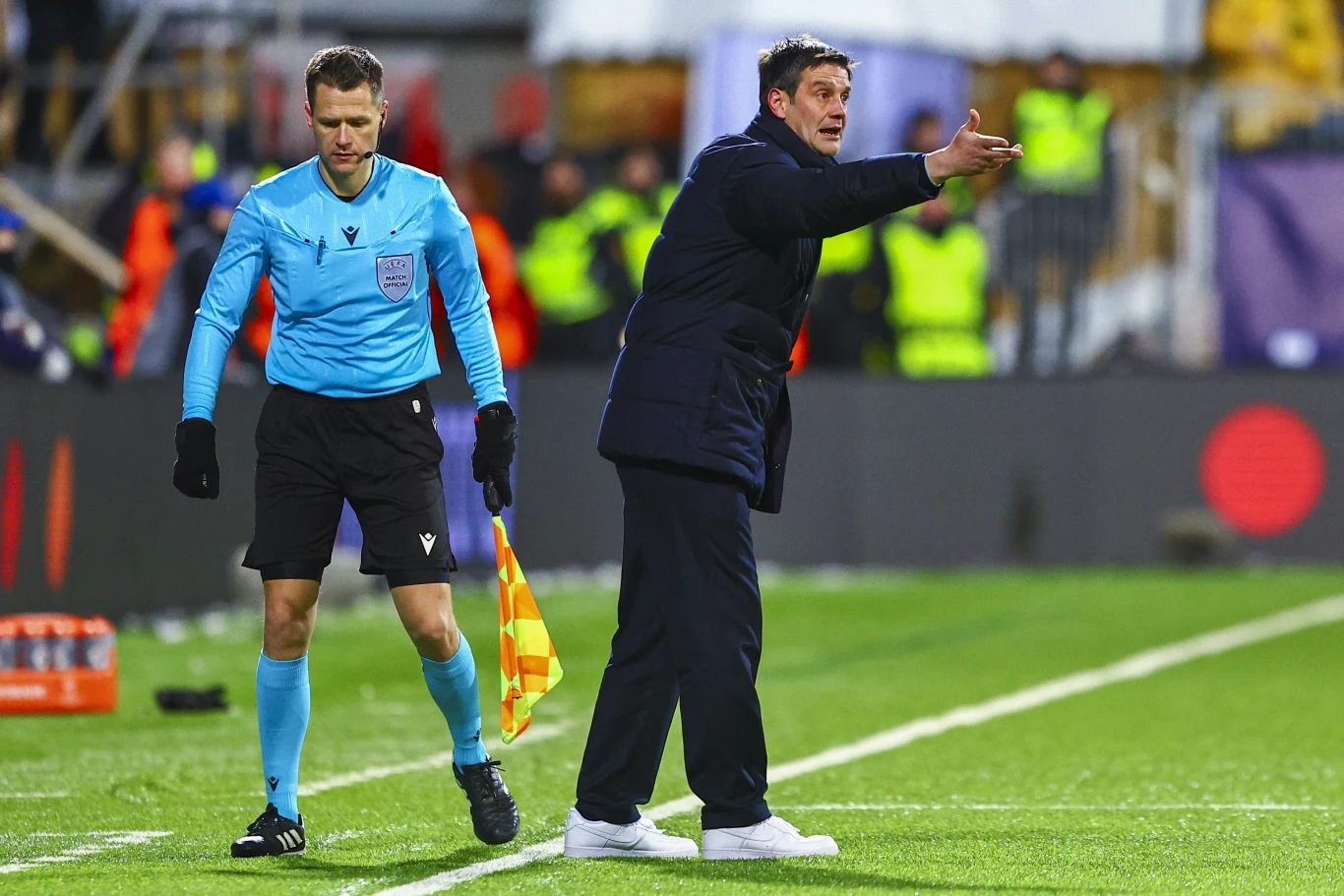 Trener w ciemnym płaszczu gestykuluje energicznie w stronę boiska, obok niego stoi sędzia boczny z chorągiewką, a w tle widoczni są stewardzi i publiczność.