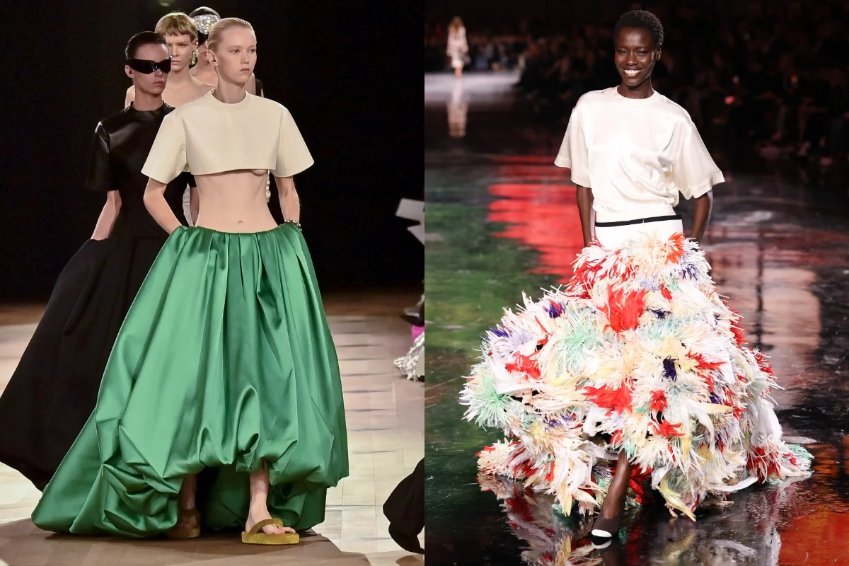 Modelki prezentujące artystyczne suknie na wybiegu, po lewej strona obszerny zielony dół i krótki biały top, po prawej spódnica ozdobiona kolorowymi piórami oraz biała bluzka, tło w ciemnych barwach z oświetlonym wybiegiem.