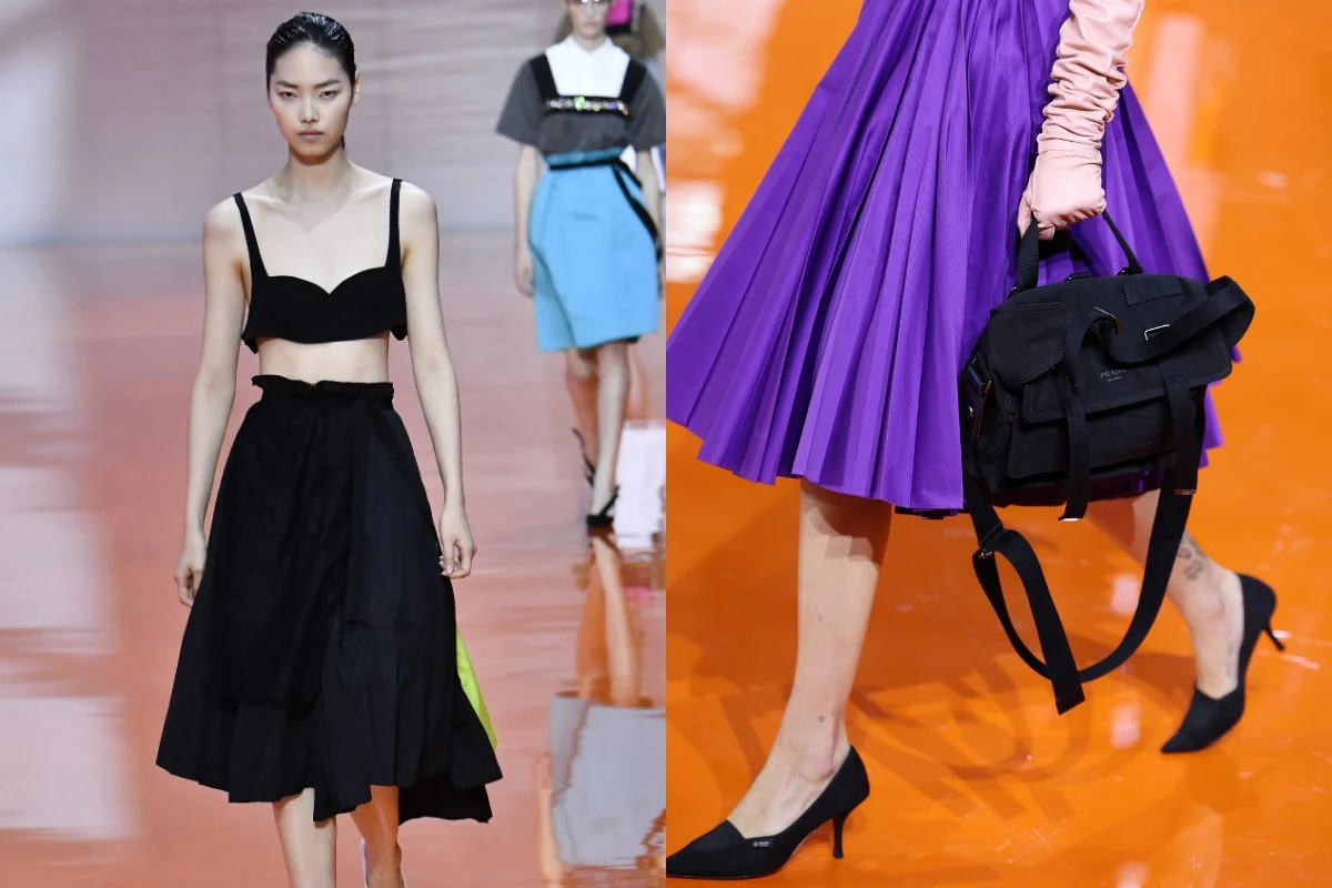 Modelki w nowoczesnych, modnych stylizacjach podczas pokazu mody na wybiegu, jedna w czarnym topie i rozkloszowanej czarnej spódnicy, druga w fioletowej spódnicy, różowej bluzce i z czarną torbą oraz czarnych szpilkach, całość na pomarańczowym tle wybi...