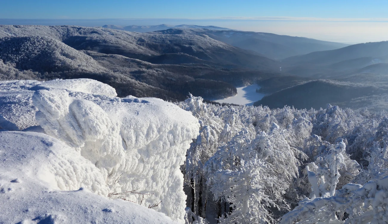 Sninský kameň w górach Vihorlat Gruba warstwa śniegu i lodu pokrywająca klif oraz drzewa, rozległa panorama gór, w oddali zamarznięte jezioro i delikatna mgła w dolinach pod błękitnym niebem.