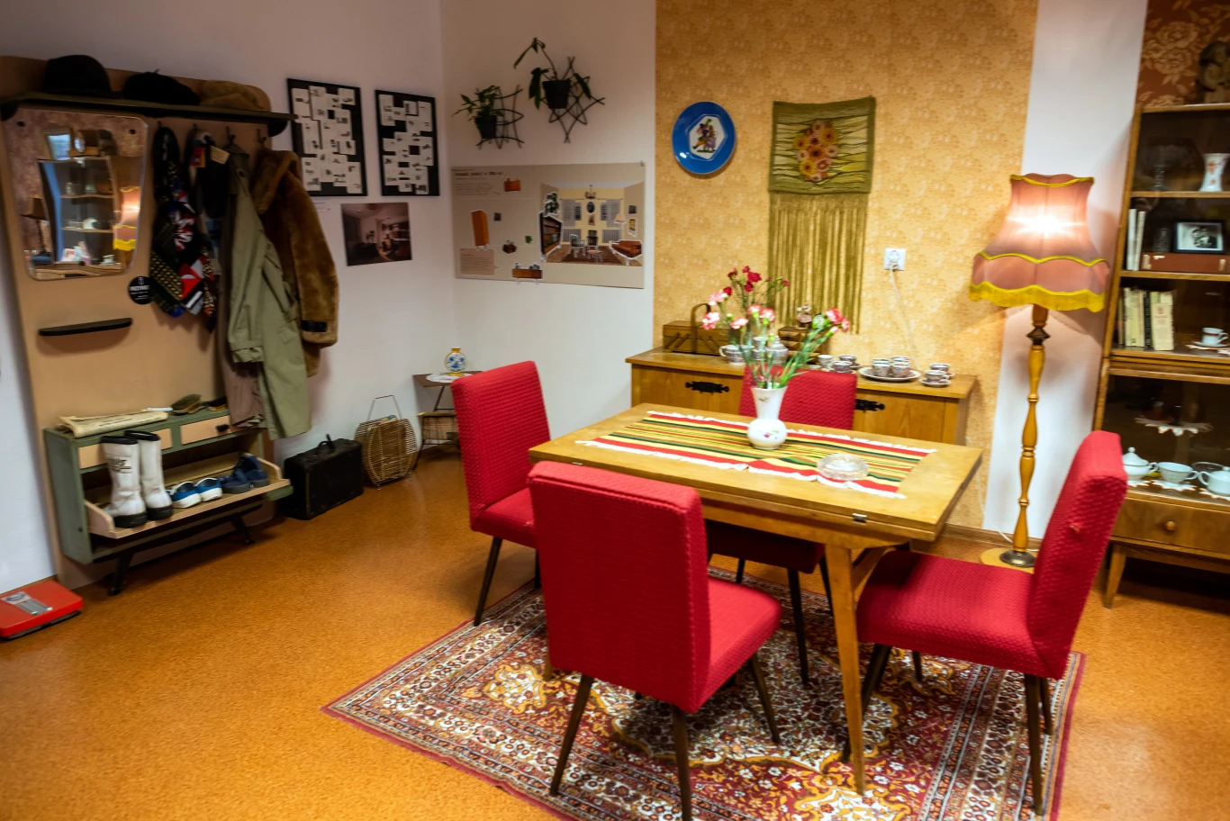 Pokój urządzony w stylu retro z drewnianym stołem, czterema czerwonymi krzesłami, dywanem i lampą stojącą, ściany udekorowane zdjęciami i ozdobami, w pokoju widoczne także kredens, wieszak z płaszczami i różne drobiazgi.