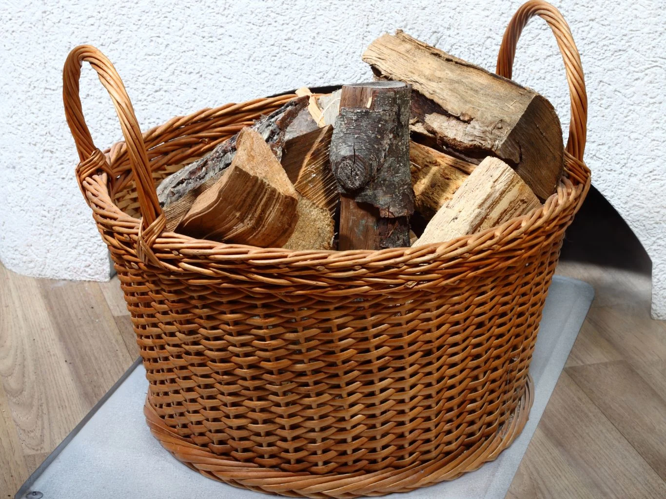 Wiklinowy kosz wypełniony pociętymi kawałkami drewna opałowego, ustawiony na jasnej podłodze przy ścianie.