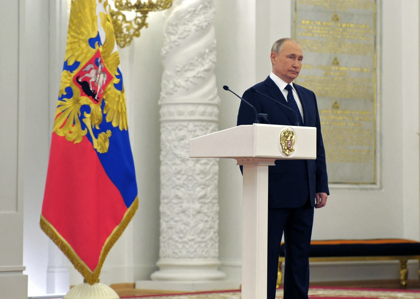 Postać mężczyzny w garniturze stojącego przy mównicy ozdobionej herbem, w tle widoczna duża flaga państwowa z godłem oraz eleganckie wnętrze o bogatej dekoracji.