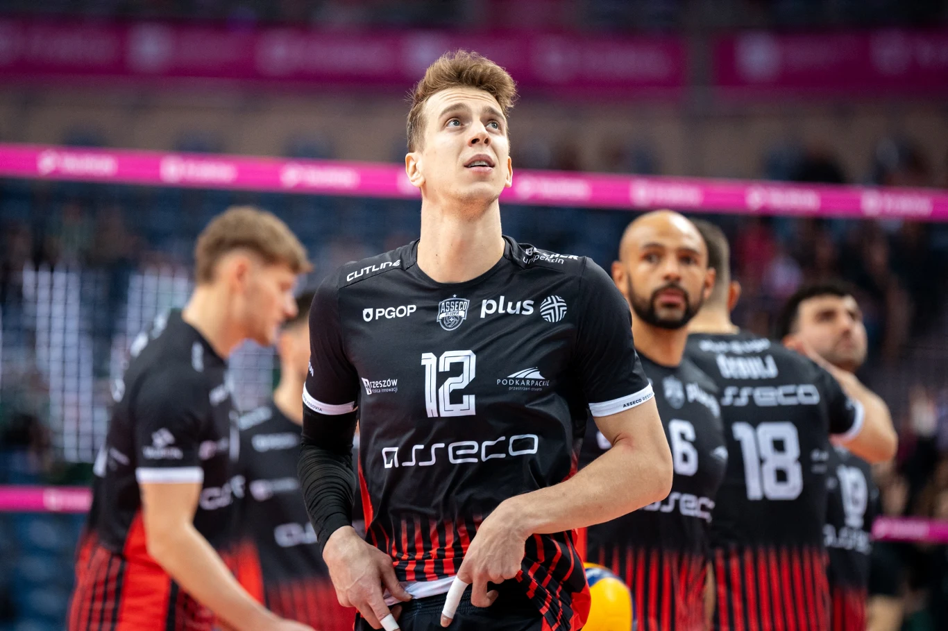 Siatkarz w czarno-czerwonym stroju z numerem 12 na piersi stoi na boisku, wokół niego widać innych członków drużyny w podobnych strojach, w tle rozmazana widownia i hala sportowa.