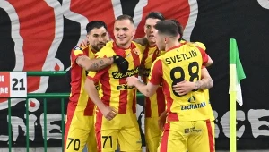 Grupa piłkarzy w żółto-czerwonych strojach świętuje zdobycie gola przy narożniku boiska, otoczona przez baner z nazwą klubu i zielone barierki.