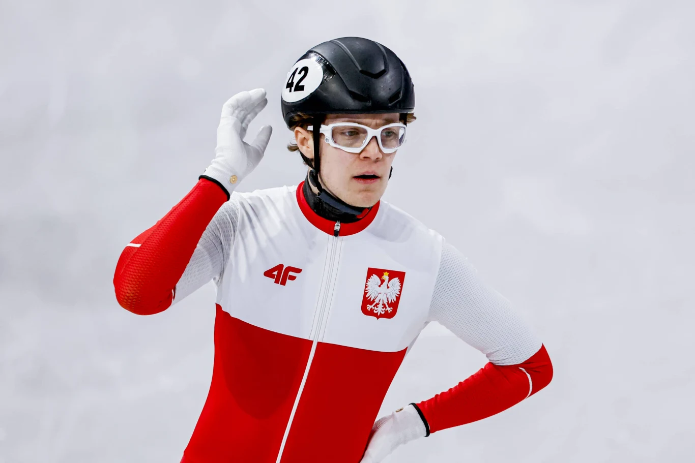 Felix Pigeon Zawodnik w stroju polskiej reprezentacji narodowej na łyżwach, ubrany w biało-czerwony kombinezon z orłem na piersi, kask i okulary ochronne, na tle śnieżnego toru.