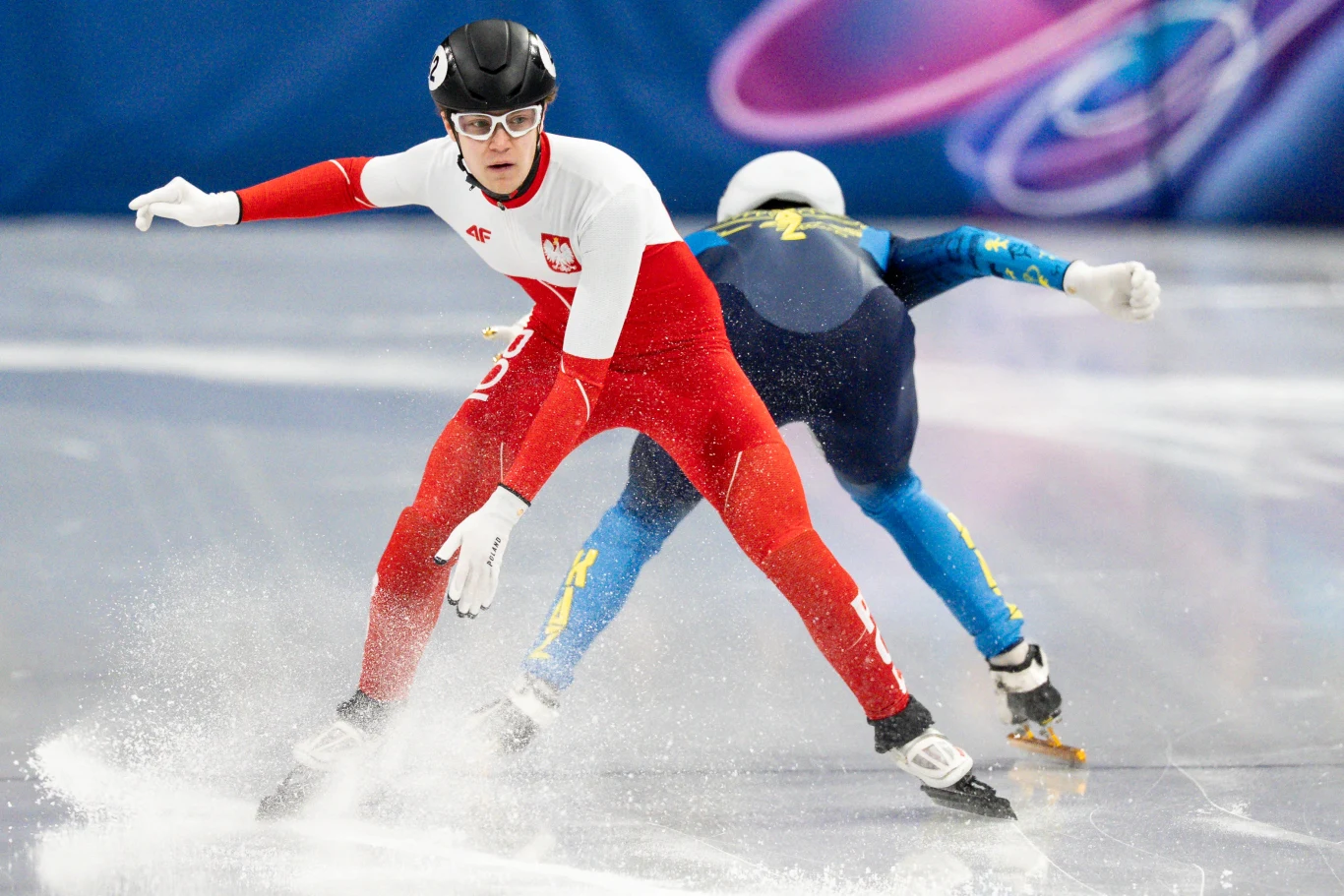 Felix Pigeon Dwoje łyżwiarzy szybkich na torze lodowym, sportowiec na pierwszym planie w biało-czerwonym stroju z polskim godłem, dynamiczna pozycja podczas skrętu, widoczne rozpryskujące się kawałki lodu.