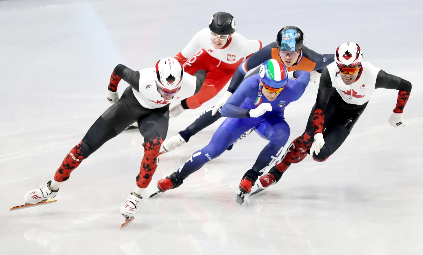 Felix Pigeon w trakcie rywalizacji w short tracku Zawodnicy w barwnych strojach w trakcie wyścigu na lodowisku, kilku panczenistów pokonujących zakręt z szybkością, koncentracją i dynamiką, widoczne narodowe emblematy na ich kombinezonach.