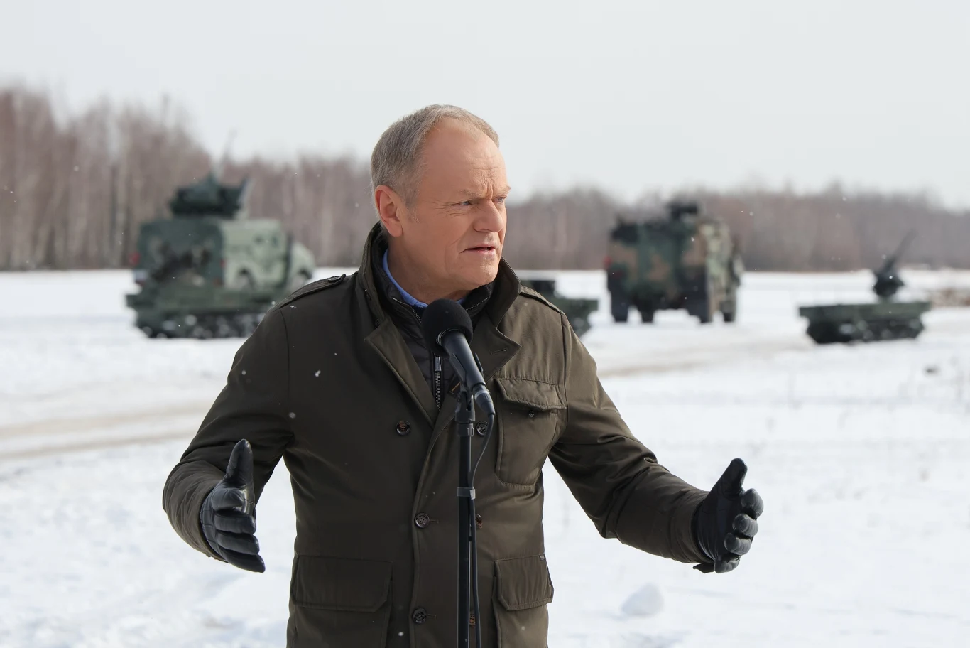 Premier Donald Tusk Mężczyzna przemawiający do mikrofonu na zaśnieżonym terenie, w tle znajdują się wojskowe pojazdy opancerzone na tle zimowego lasu.