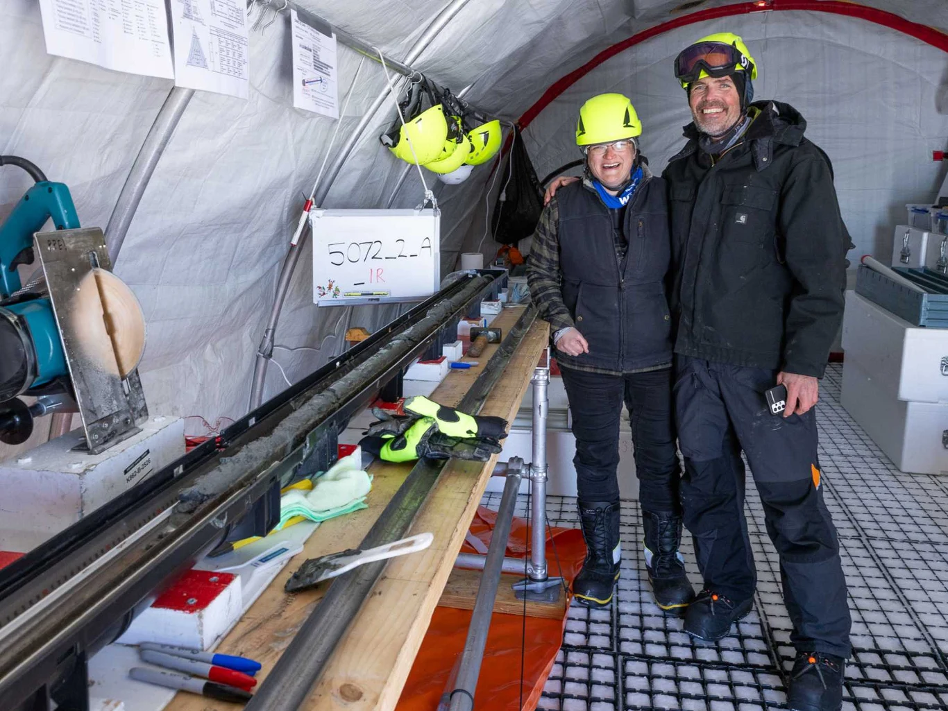 Współliderzy SWAIS2C Molly Paterson i Huw Horgan z pierwszym wydobytym fragmentem rdzenia Dwójka naukowców ubranych w ciepłe ubrania i żółte kaski stoi w namiocie laboratoryjnym, obok długiego rdzenia lodowego ułożonego na stole roboczym. W tle widoczne są narzędzia, kaski oraz różne przedmioty laboratoryjne.
