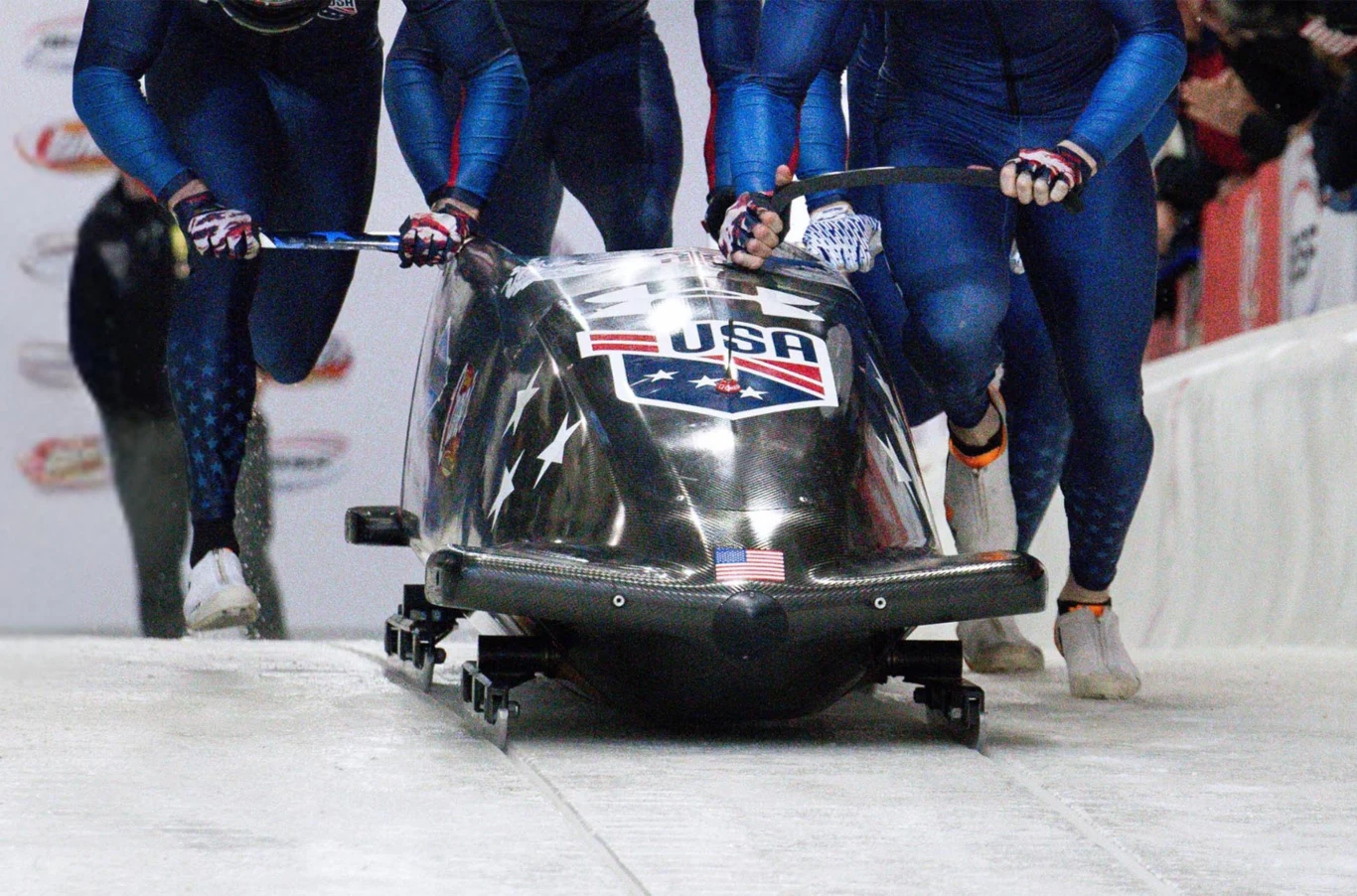 Bobsleje Team USA w tunelu aerodynamicznym Hondy. Czterech sportowców w sportowych kombinezonach rozpędza bobslej z napisem USA na lodowym torze, widać dynamiczny moment startu zawodów.