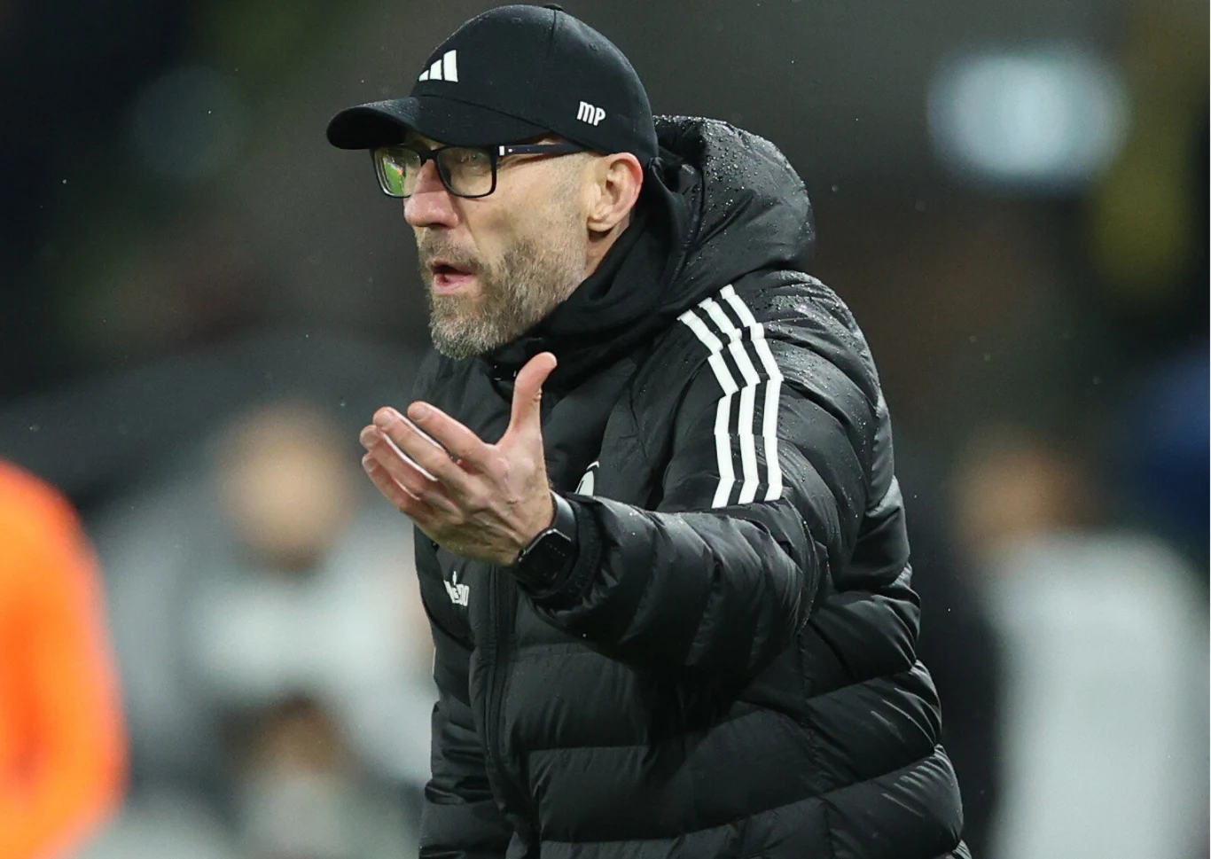 Mężczyzna w sportowej kurtce i czapce z daszkiem wykonuje gest ręką podczas wydarzenia sportowego, wygląda na zaangażowanego i skoncentrowanego na sytuacji na boisku.