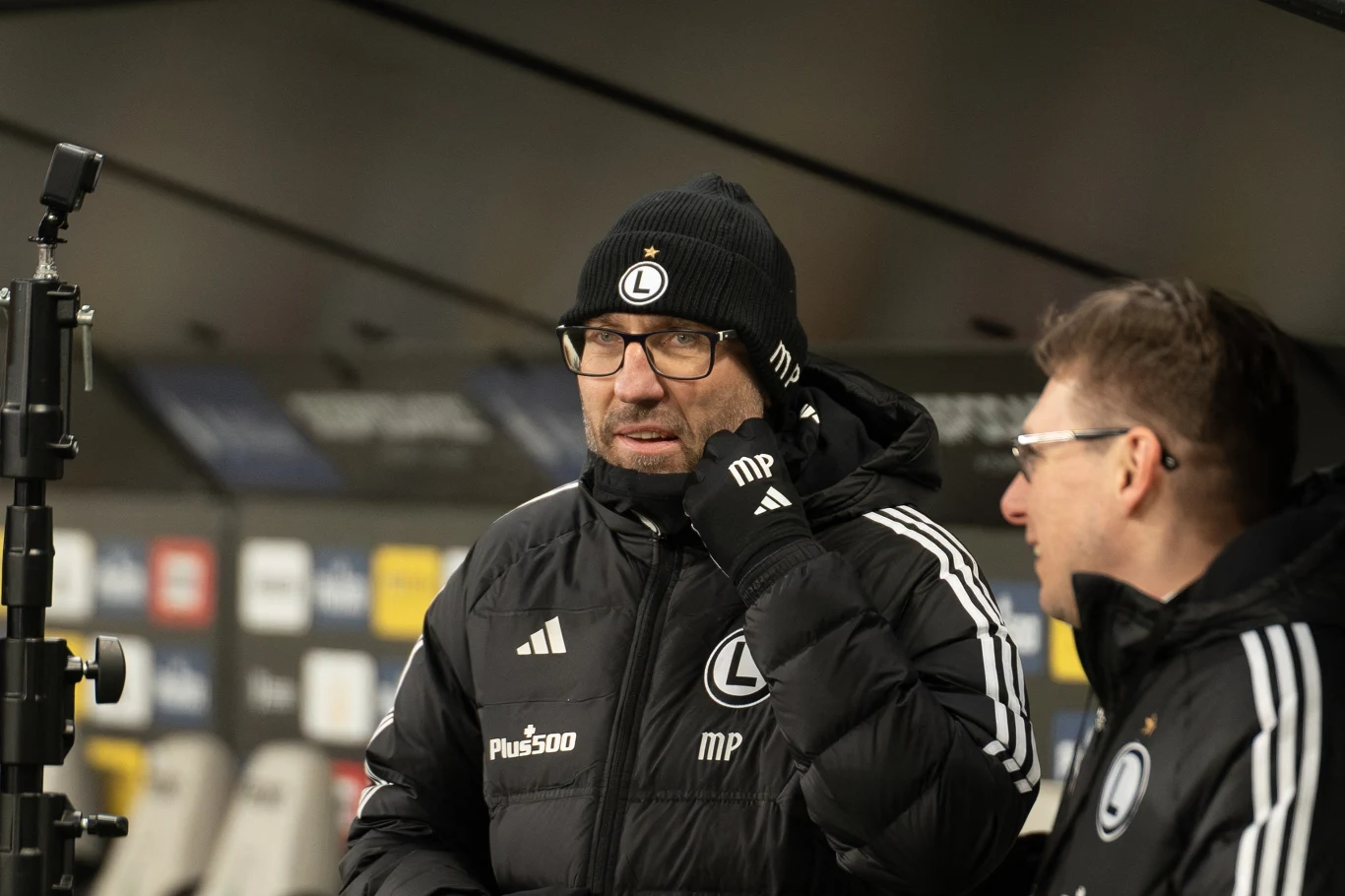 Dwóch mężczyzn w sportowych kurtkach i czapkach z logo drużyny piłkarskiej Legia Warszawa stoi na stadionowej ławce rezerwowych, jeden z nich ma brodę i okulary, drugi rozmawia z nim, w tle rozmyte tablice sponsorskie.