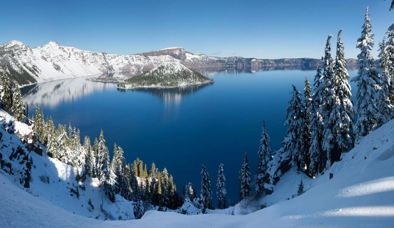 Crater Lake to jedno z najczystszych jezior na świecie Błękitne jezioro otoczone stromymi, ośnieżonymi zboczami gór, z wyłaniającą się z wody wyspą, przy brzegu rosną gęsto pokryte śniegiem drzewa iglaste, wszystko pod czystym, niebieskim niebem.