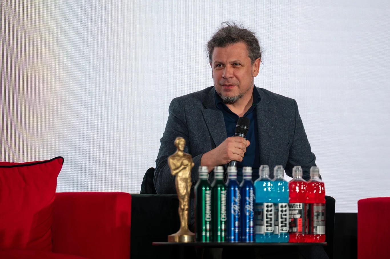 Mężczyzna w marynarce trzyma mikrofon i siedzi na czerwonej sofie, przed nim znajduje się statuetka przypominająca Oscara oraz kilka butelek napojów izotonicznych w różnych kolorach, tło białe minimalistyczne.