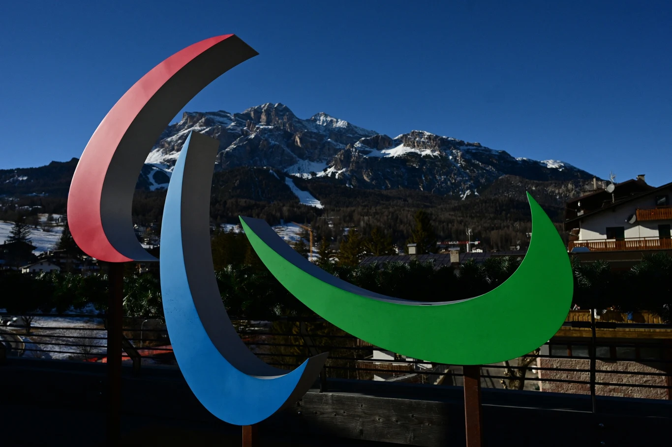 Logo igrzysk paraolimpijskich na tle ośnieżonych gór i zabudowań górskiego miasteczka, w słoneczny dzień.