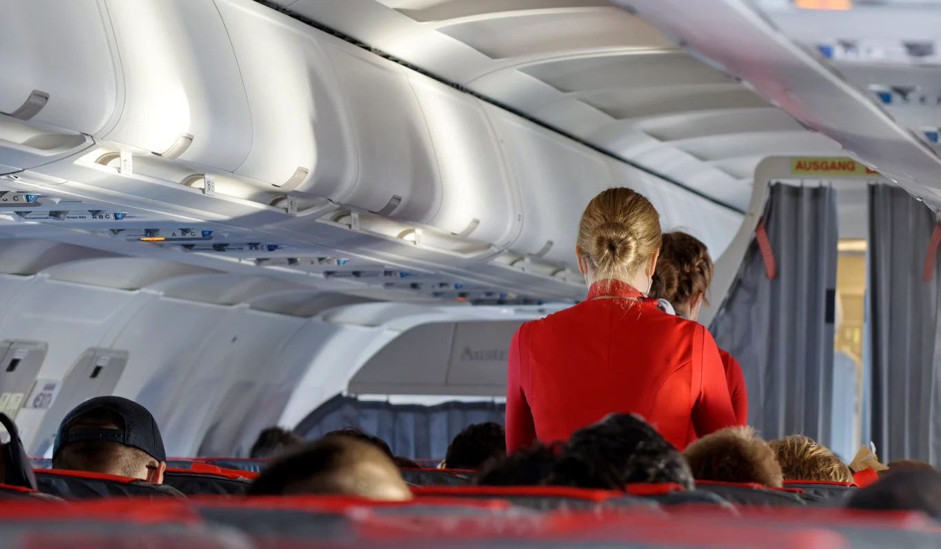 Dziennikarza AFP nie wpuszczono na pokład samolotu, zdj. ilustracyjne Wnętrze samolotu pasażerskiego z widocznymi pasażerami siedzącymi w fotelach oraz stewardesą w czerwonym uniformie obsługującą podróżnych.