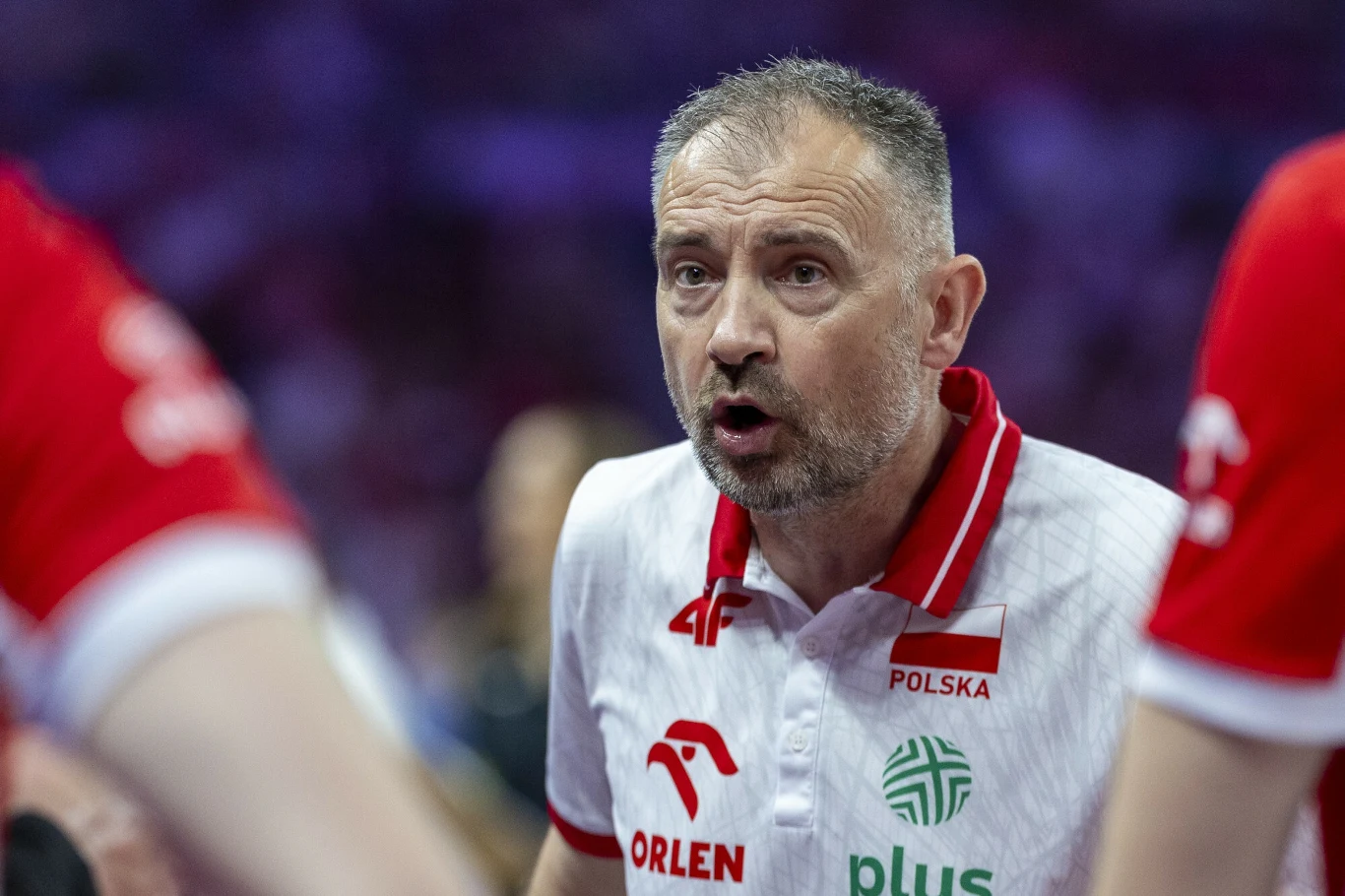 Nikola Grbić Trener drużyny siatkarskiej Polski w białej koszulce z logotypami sponsorów, wyraża emocje podczas rozmowy z zawodnikami, w tle rozmyte postacie innych członków drużyny.