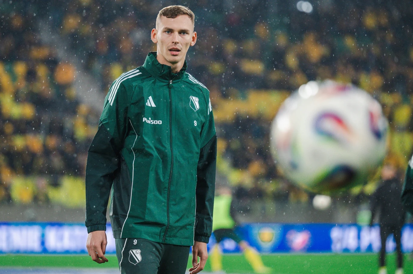 Młody mężczyzna w zielonej kurtce sportowej z logo klubu piłkarskiego stoi na boisku, w tle rozmyte trybuny z kibicami w żółtych ubraniach, na pierwszym planie piłka w locie.