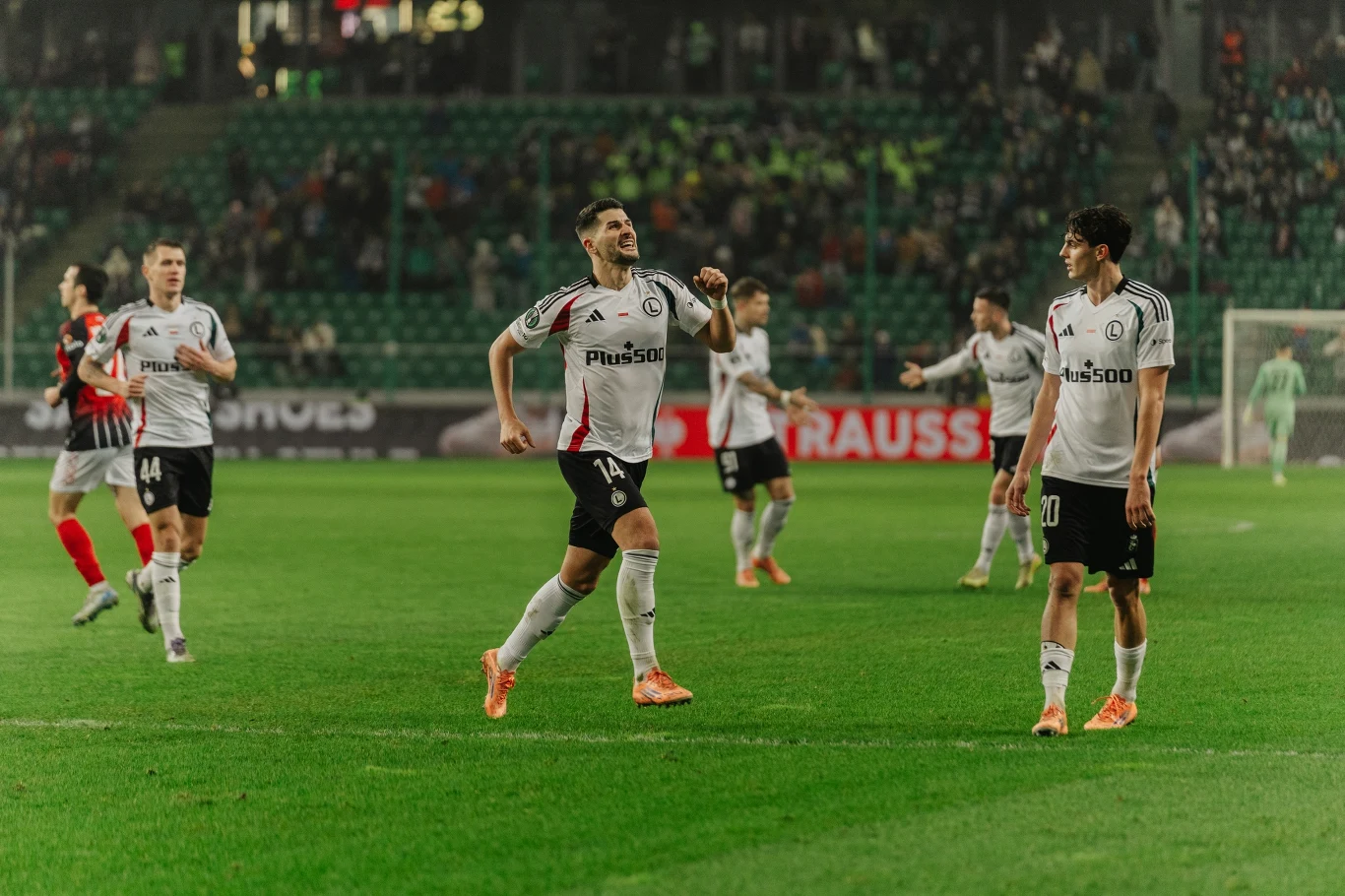 Piłkarze w białych i czerwonych strojach rywalizują na boisku podczas meczu piłki nożnej, na trybunach widoczna publiczność, a zawodnik z uniesioną ręką świętuje udaną akcję.