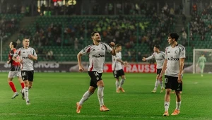 Piłkarze w białych i czerwonych strojach rywalizują na boisku podczas meczu piłki nożnej, na trybunach widoczna publiczność, a zawodnik z uniesioną ręką świętuje udaną akcję.