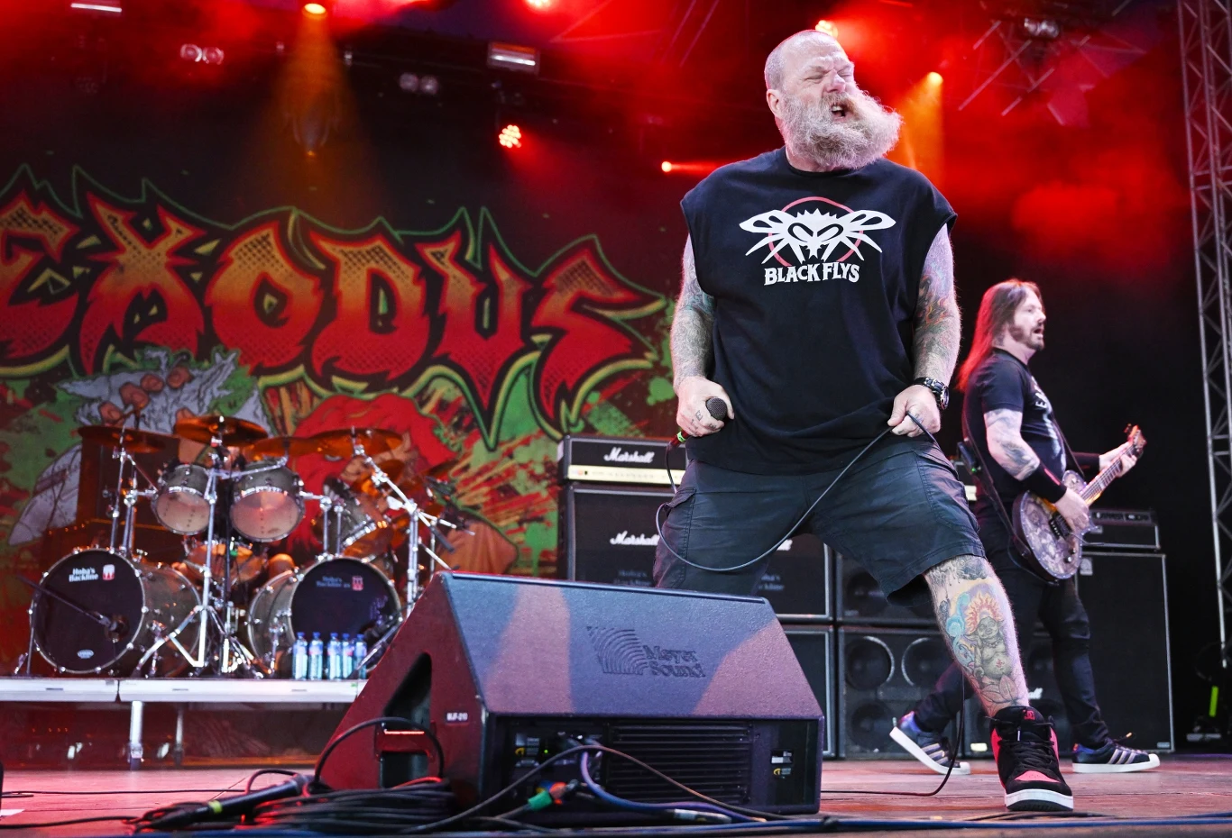 Rob Dukes (Exodus) w akcji Zespół muzyczny podczas występu na scenie, kolorowe światła i charakterystyczne logo zespołu w tle, wokalista z wyrazistą brodą oraz tatuażami na rękach, obok gitarzysta przygotowujący się do gry, a na scenie widoczne są instrumenty muzyczne i sprzęt n...