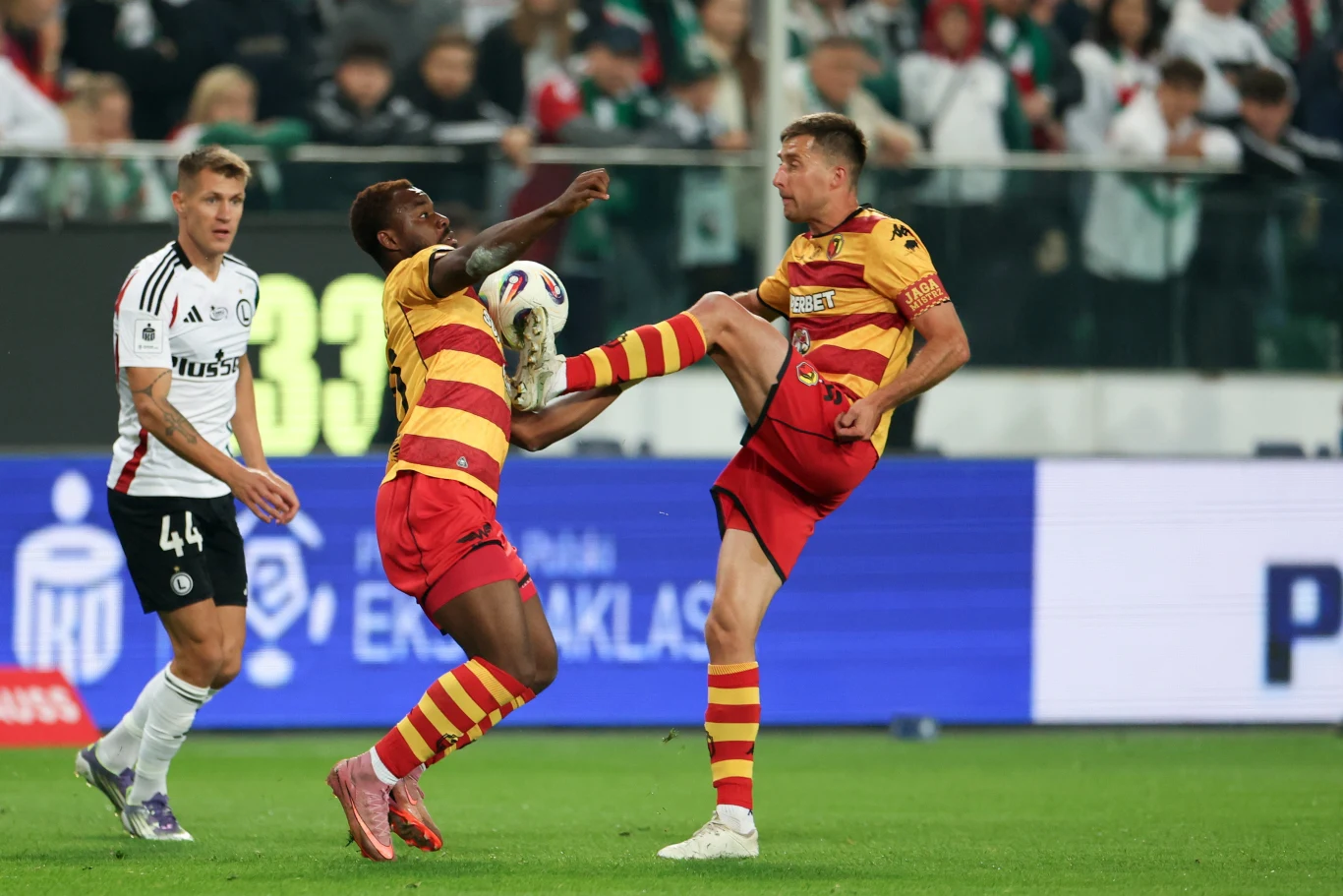 Jagiellonia zagra osłabiona brakiem Afimico Pululu i Tarasa Romanczuka Dwóch piłkarzy w żółto-czerwonych strojach walczy o piłkę, podnosząc nogi na wysokość tułowia, w tle widoczny zawodnik w białej koszulce oraz kibice na trybunach.