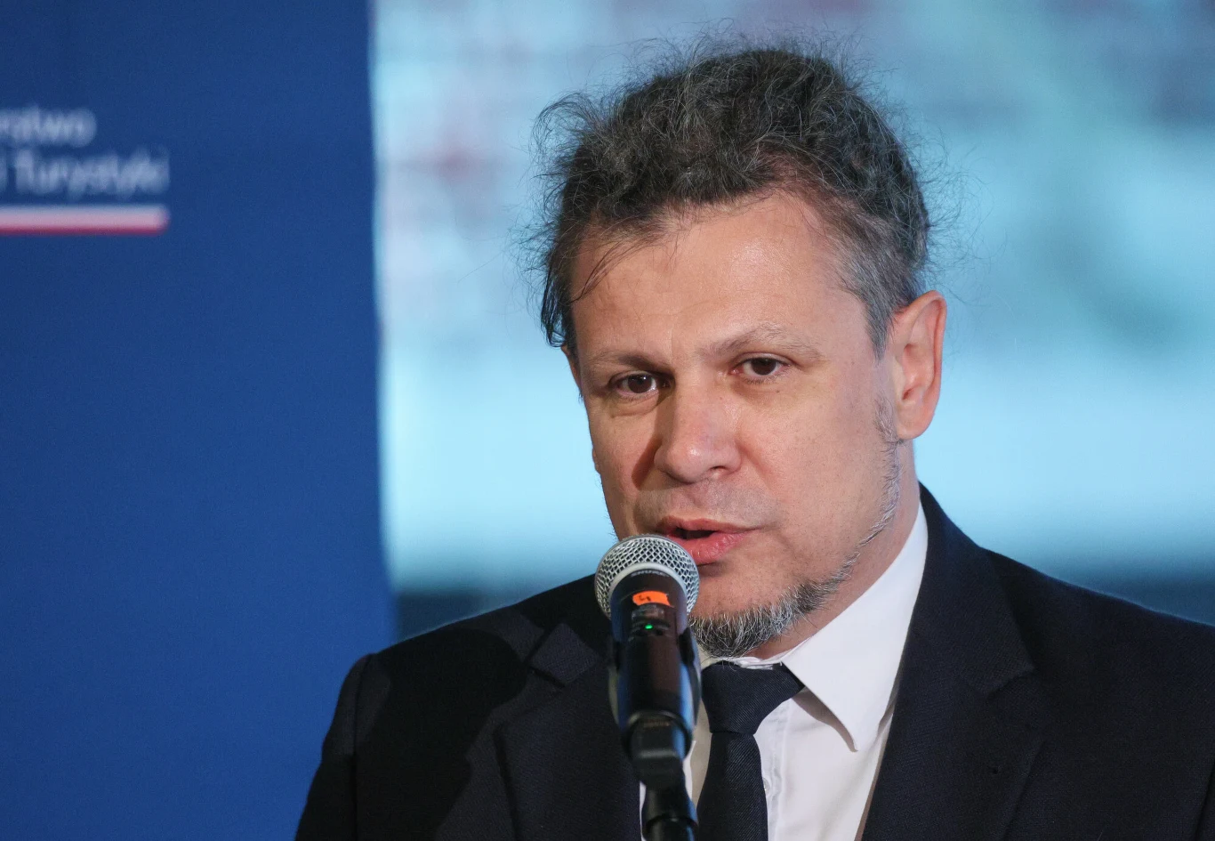 Mężczyzna w średnim wieku przemawia do mikrofonu, ujęty na tle niebieskiego baneru z logo instytucji.