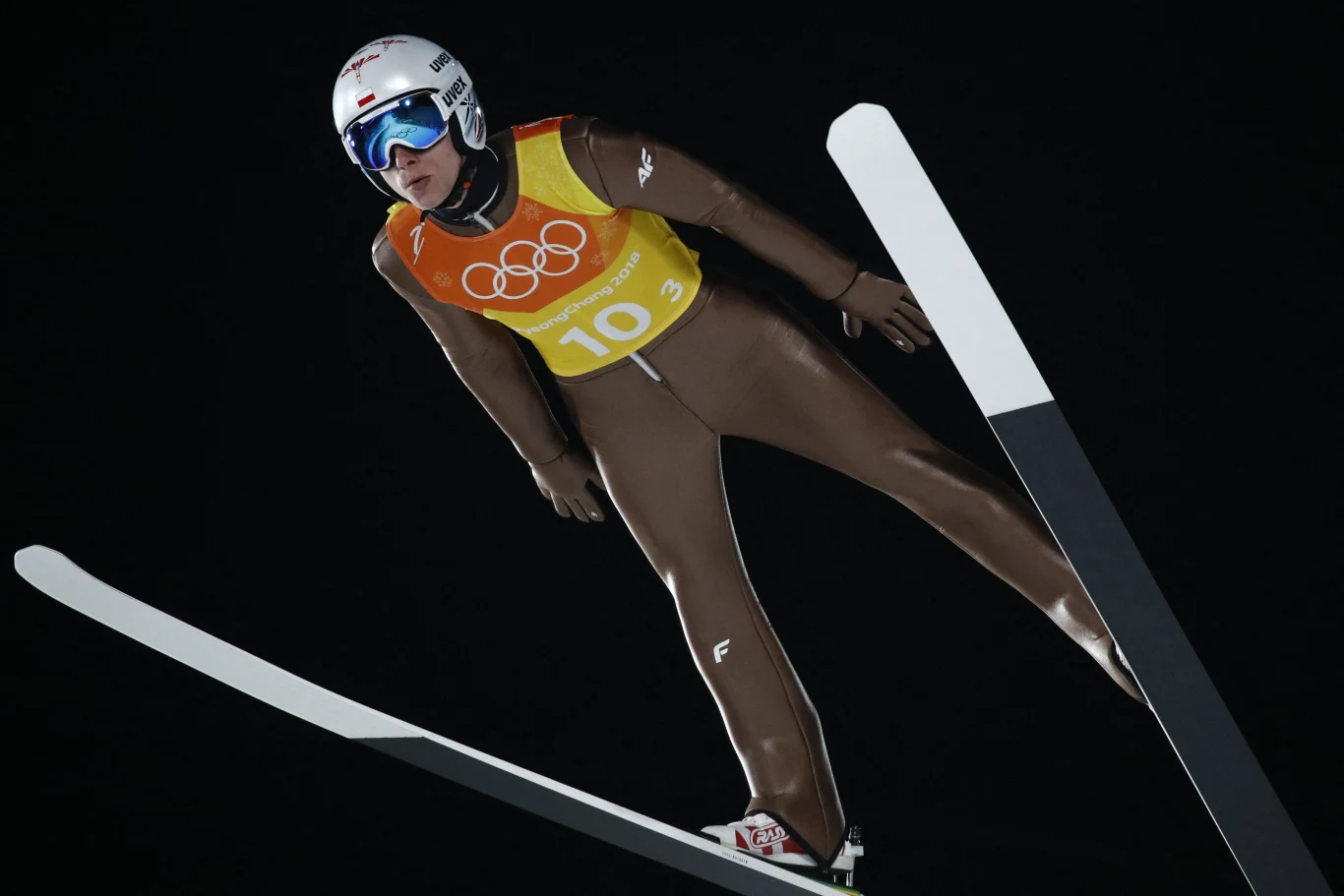 Zawodnik w brązowym kombinezonie i kasku w trakcie skoku narciarskiego w powietrzu, na tle nocnego nieba, z widocznym numerem startowym oraz symbolem igrzysk olimpijskich na kamizelce.