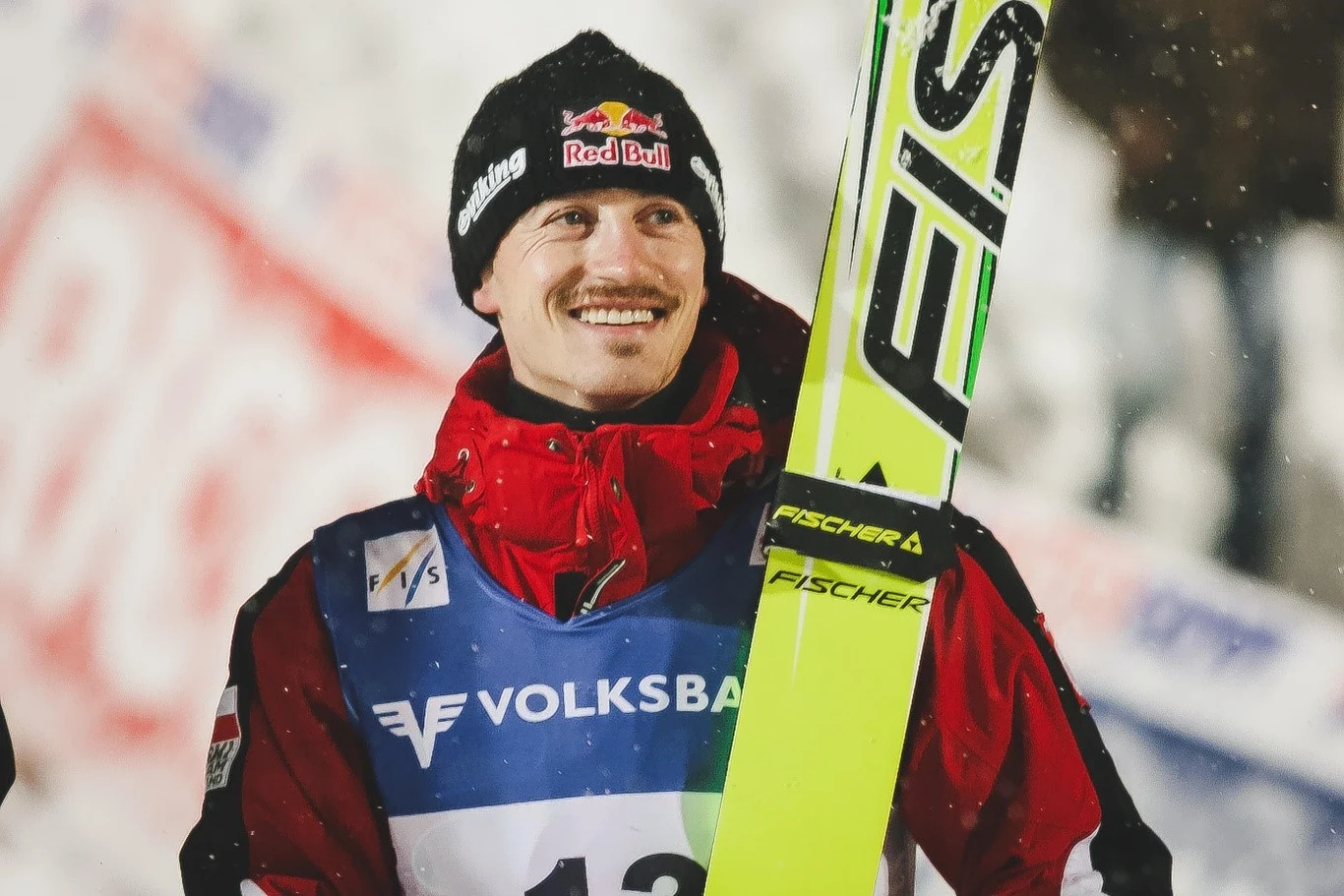 Uśmiechnięty skoczek narciarski w czerwonej kurtce z numerem startowym trzyma narty, w tle rozmyte elementy trybun i banerów.