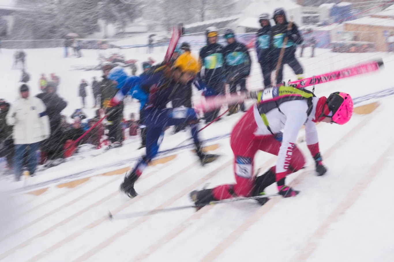 Narciarze upadający podczas rywalizacji na śnieżnym stoku, w tle widoczna publiczność oraz sędziowie uważnie obserwujący przebieg zawodów.