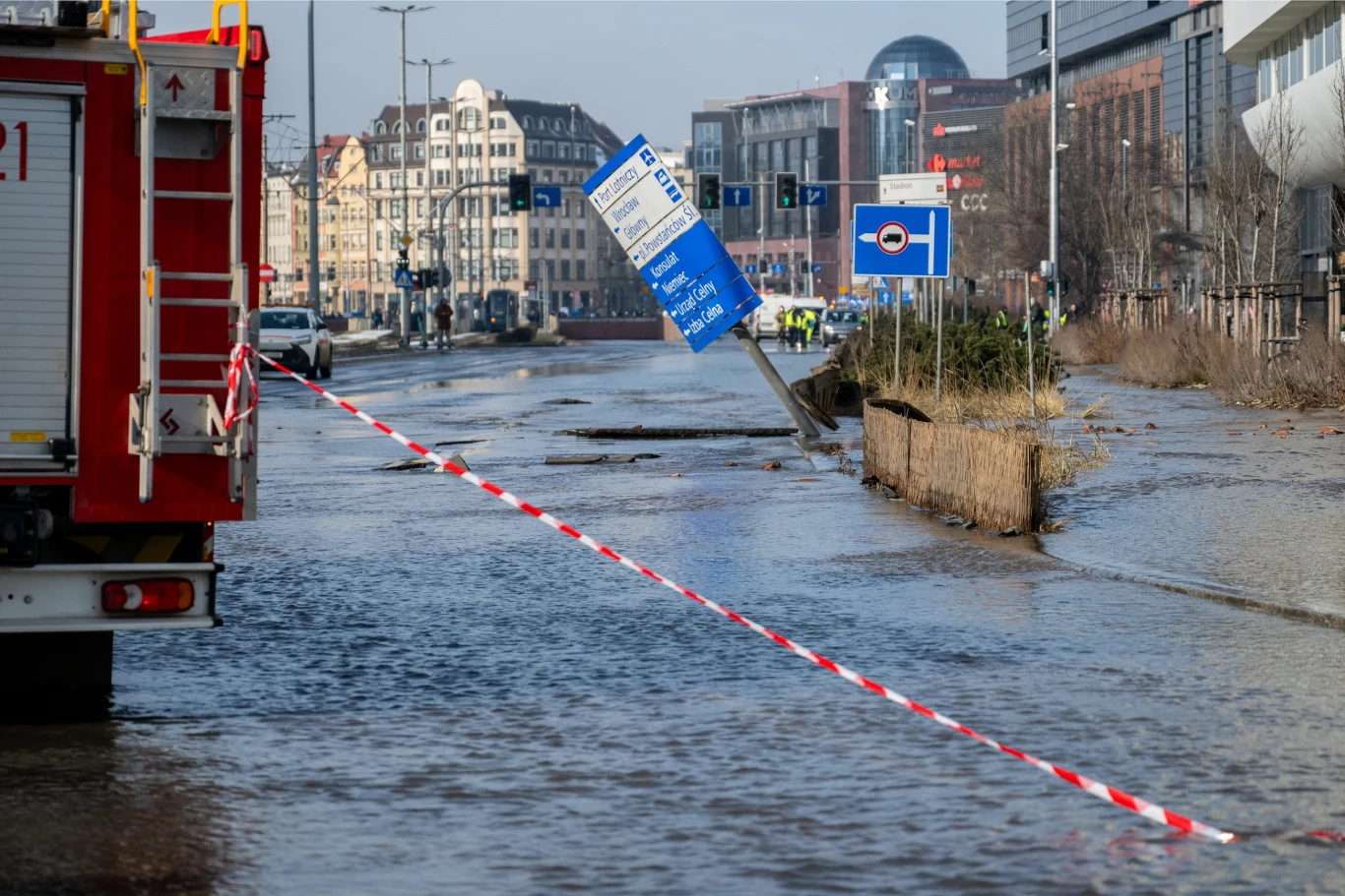 Zalana ulica w centrum miasta, widoczny samochód strażacki po lewej stronie, przewrócona tablica informacyjna oraz taśma zabezpieczająca teren. W tle budynki oraz kilka pojazdów służb ratunkowych.