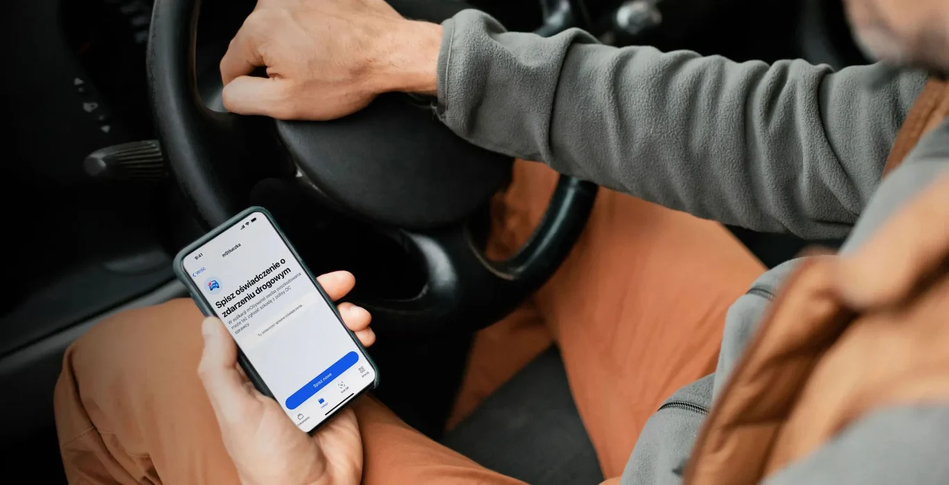 Mężczyzna w kurtce i spodniach w kolorze camel prowadzi samochód, trzymając w jednej ręce kierownicę, a w drugiej smartfon z uruchomioną aplikacją.
