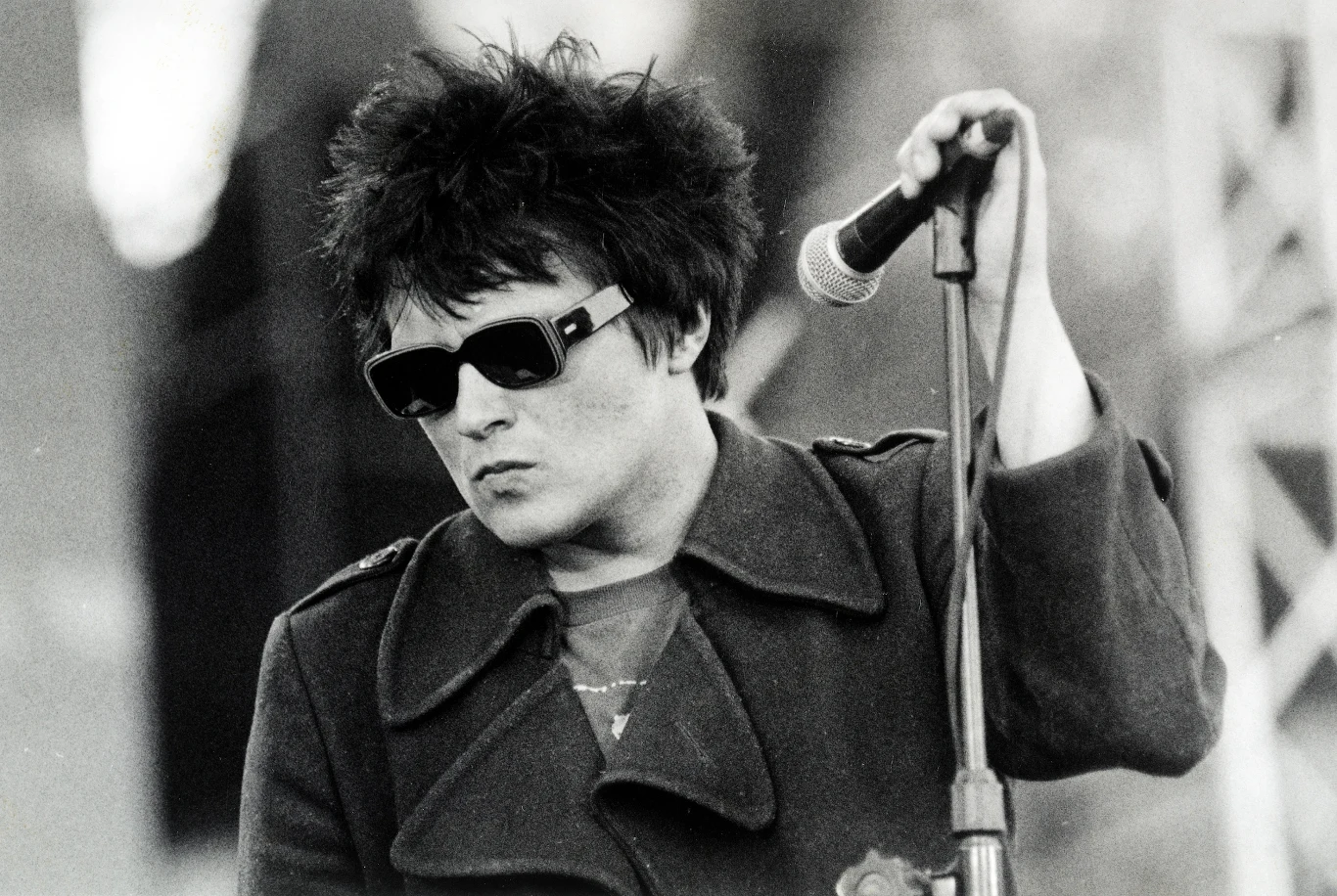 Muniek Staszczyk (T.Love) w latach 90. Mężczyzna w ciemnych okularach i płaszczu, stojący przy mikrofonie, trzymający jego statyw, z potarganymi włosami i poważnym wyrazem twarzy, na tle nieostrego, scenicznego oświetlenia.