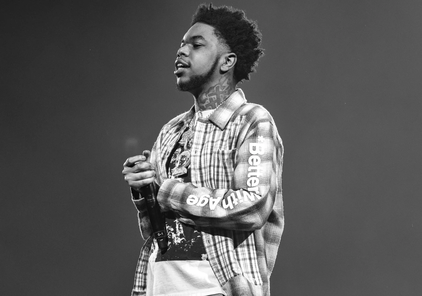 Lil Poppa zmarł w wieku 25 lat Mężczyzna o charakterystycznym wyglądzie, ubrany w koszulę w kratę z napisem na rękawie oraz koszulkę z nadrukiem, stoi na scenie, złączył dłonie, patrzy w bok, tło rozmyte i jednolite.