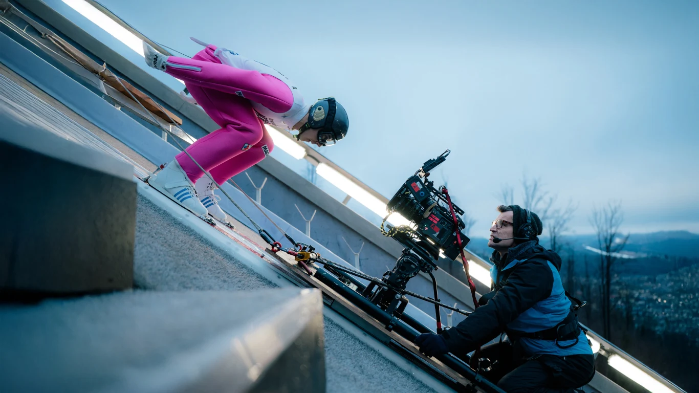 Skoczek narciarski w różowym kombinezonie przygotowuje się do startu na skoczni, obok niego operator filmuje całą scenę profesjonalną kamerą, w tle widoczne rozmazane krajobrazy górskie.