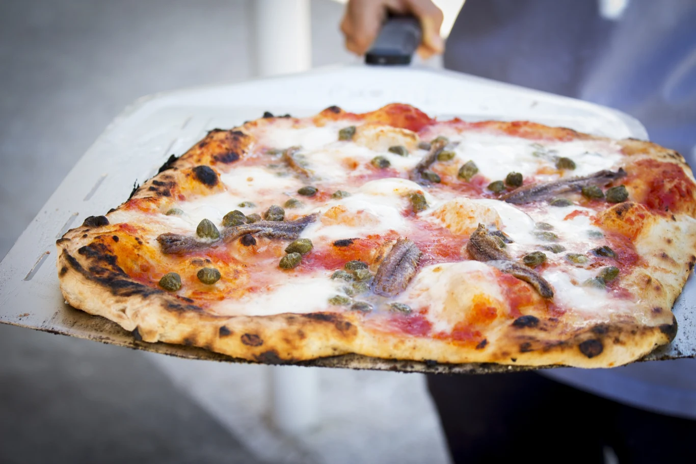 Kwadratowa pizza z widocznym sosem pomidorowym, mozzarellą, kaparami i filecikami anchois, wyjęta prosto z pieca na metalowej łopacie, z wyraźnie wypieczonymi brzegami.