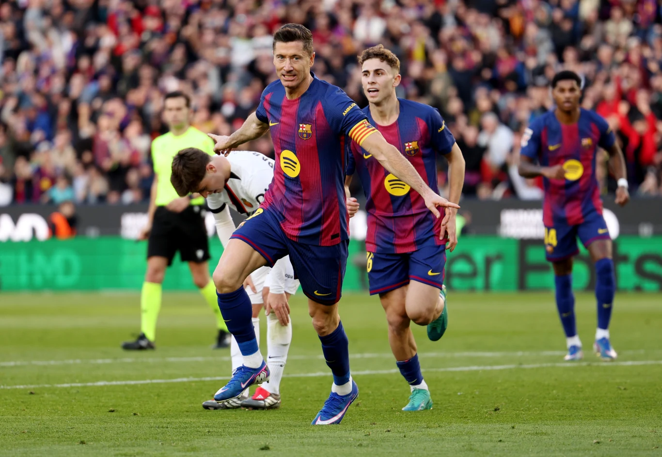 Robert Lewandowski w barwach FC Barcelona podczas meczu La Liga Piłkarze FC Barcelony świętują zdobycie gola podczas meczu piłkarskiego, na pierwszym planie zawodnik z opaską kapitańską w dynamicznej pozie, tłum kibiców na trybunach w tle.