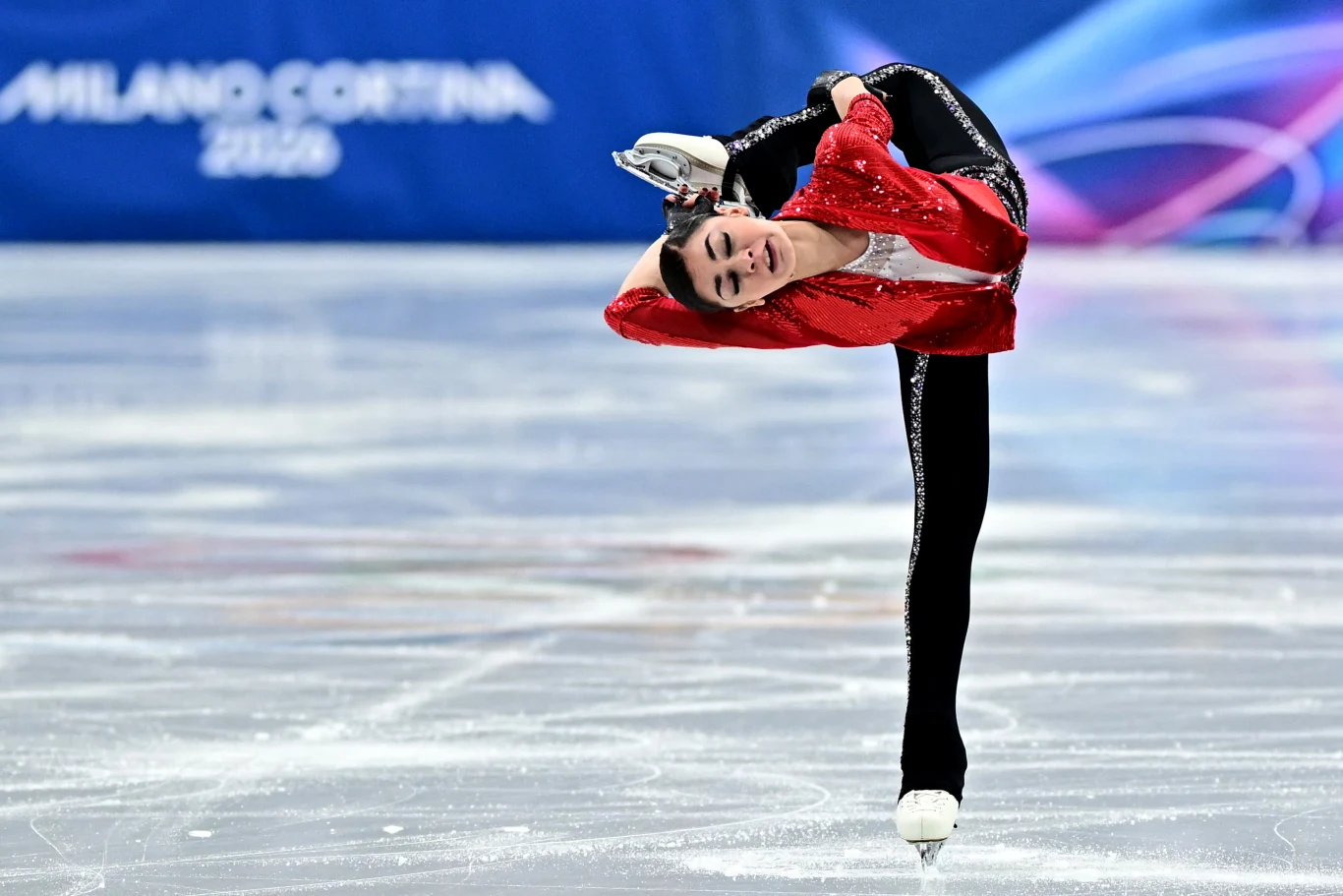 Łyżwiarka figurowa wykonuje skomplikowaną figurę na lodowisku, ubrana w czerwono-czarny strój, w tle widoczne są niebieskie dekoracje i napis Milano-Cortina 2026.