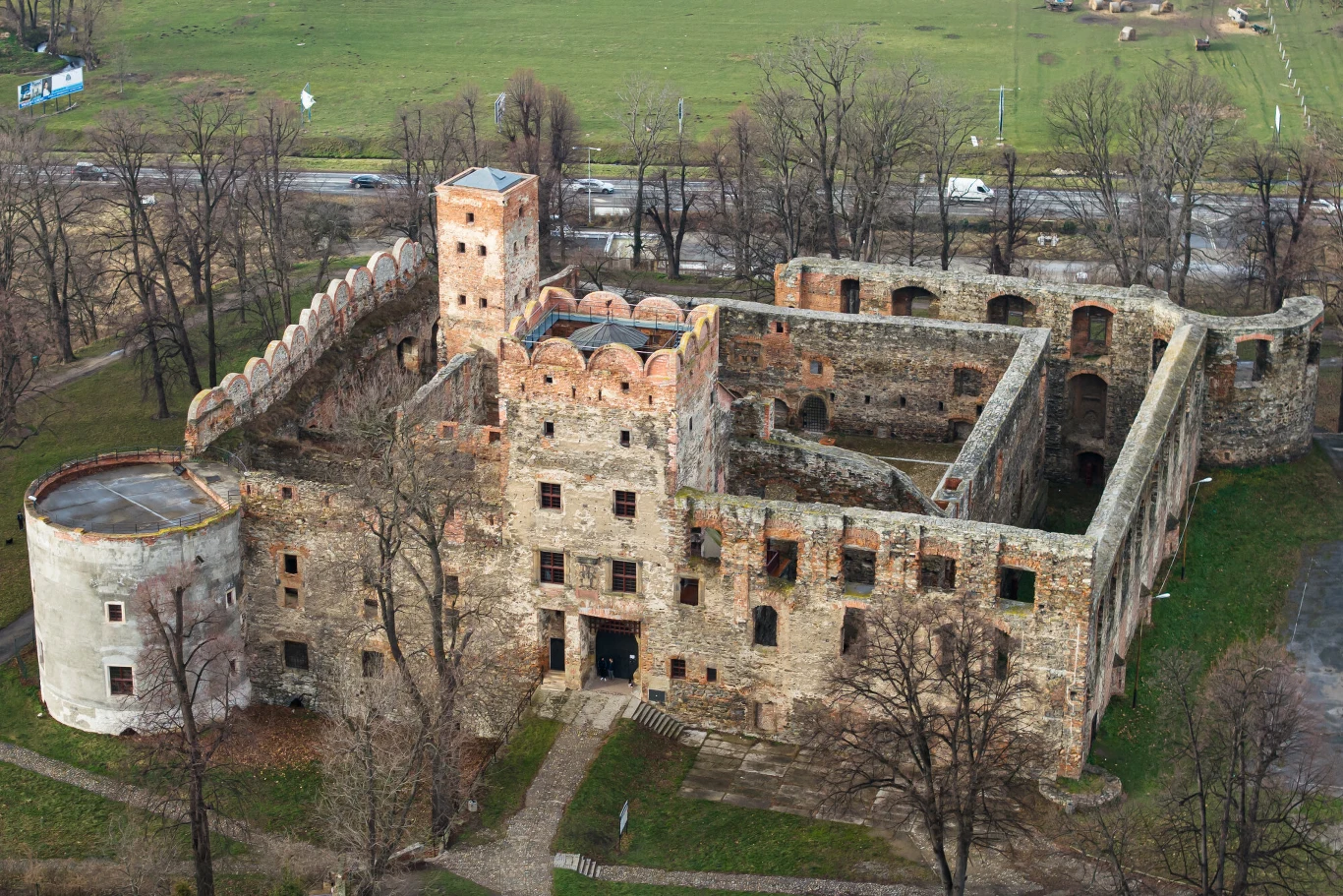 Nawet dziś Ząbkowice mogą rozbudzać zmysły miłośników historii z dreszczykiem Ruiny kamiennego zamku otoczone murami i wieżą, położone na otwartej przestrzeni z widocznymi resztkami drzew oraz drogą w tle.