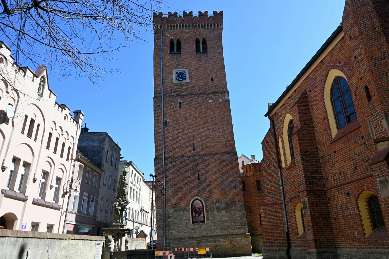 Namiastka Włoch na Dolnym Śląsku Ceglana wieża z zegarem na fasadzie otoczona zabytkowymi budynkami; obok wieży znajduje się fragment muru oraz detale architektoniczne charakterystyczne dla stylu gotyckiego.