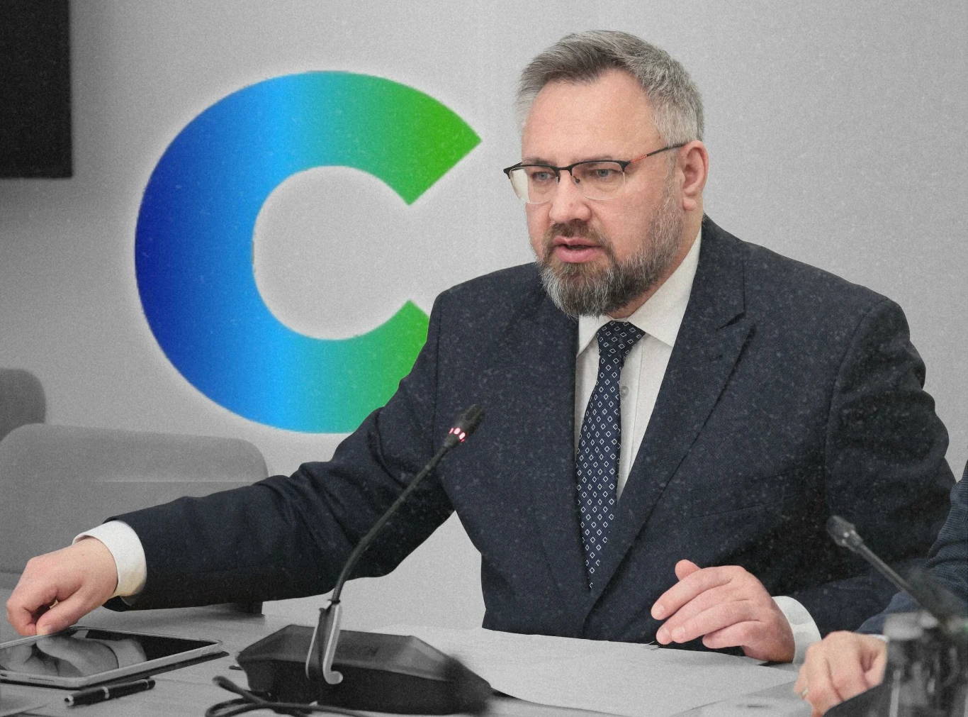 Poseł Mirosław Suchoń to najczęściej typowany kandydat na szefa klubu Centrum. Mężczyzna z brodą i okularami siedzi przy stole konferencyjnym, ubrany w garnitur i krawat, mówi do mikrofonu. W tle widoczne duże logo w odcieniach niebiesko-zielonych.