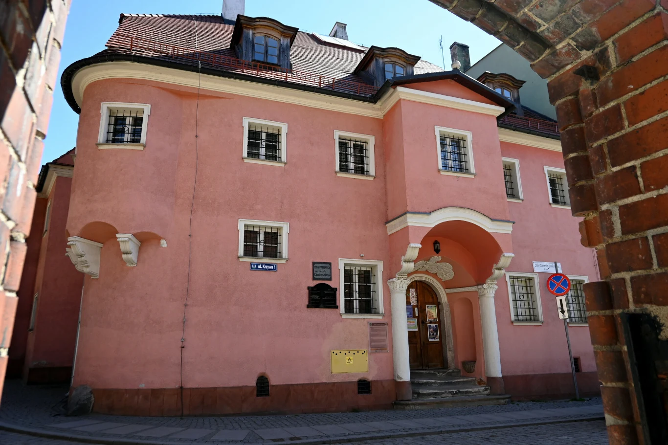 Dwór, który dziś w swoich murach skrywa niebanalne ekspozycje Różowy budynek z ozdobnym portalem wejściowym, licznymi oknami i spadzistym dachem, zlokalizowany przy brukowanej ulicy, otoczony zabytkowymi murami miejskimi. Na fasadzie widoczne tablice informacyjne oraz skrzynki pocztowe.